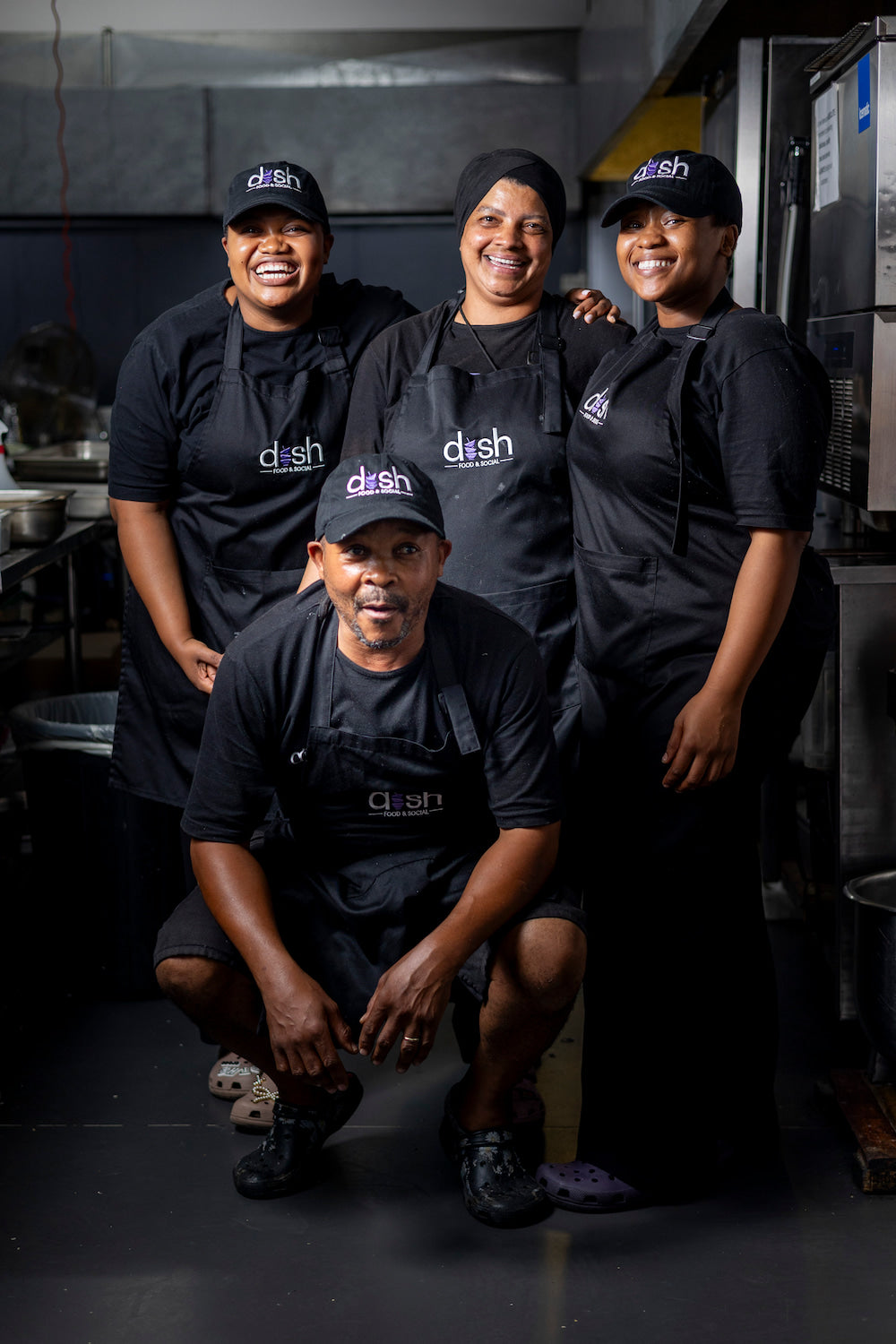 Four individuals wearing black aprons and hats with 'dish' branding in a kitchen setting.