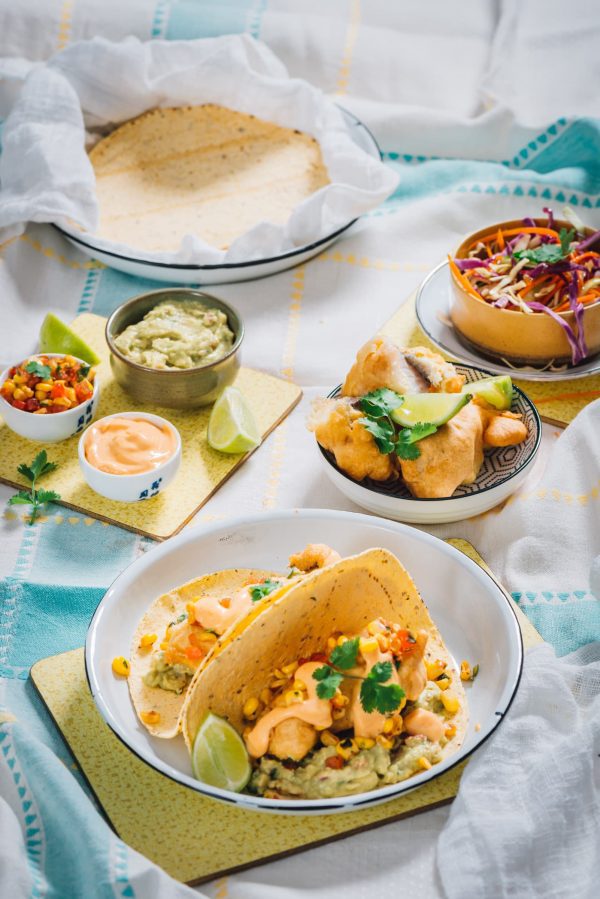 Various bowls containing crispy fried fish, corn taco shells, sriracha mayo, a white cabbage, fennel & lemon slaw, and an avo, pea & mint guacamole