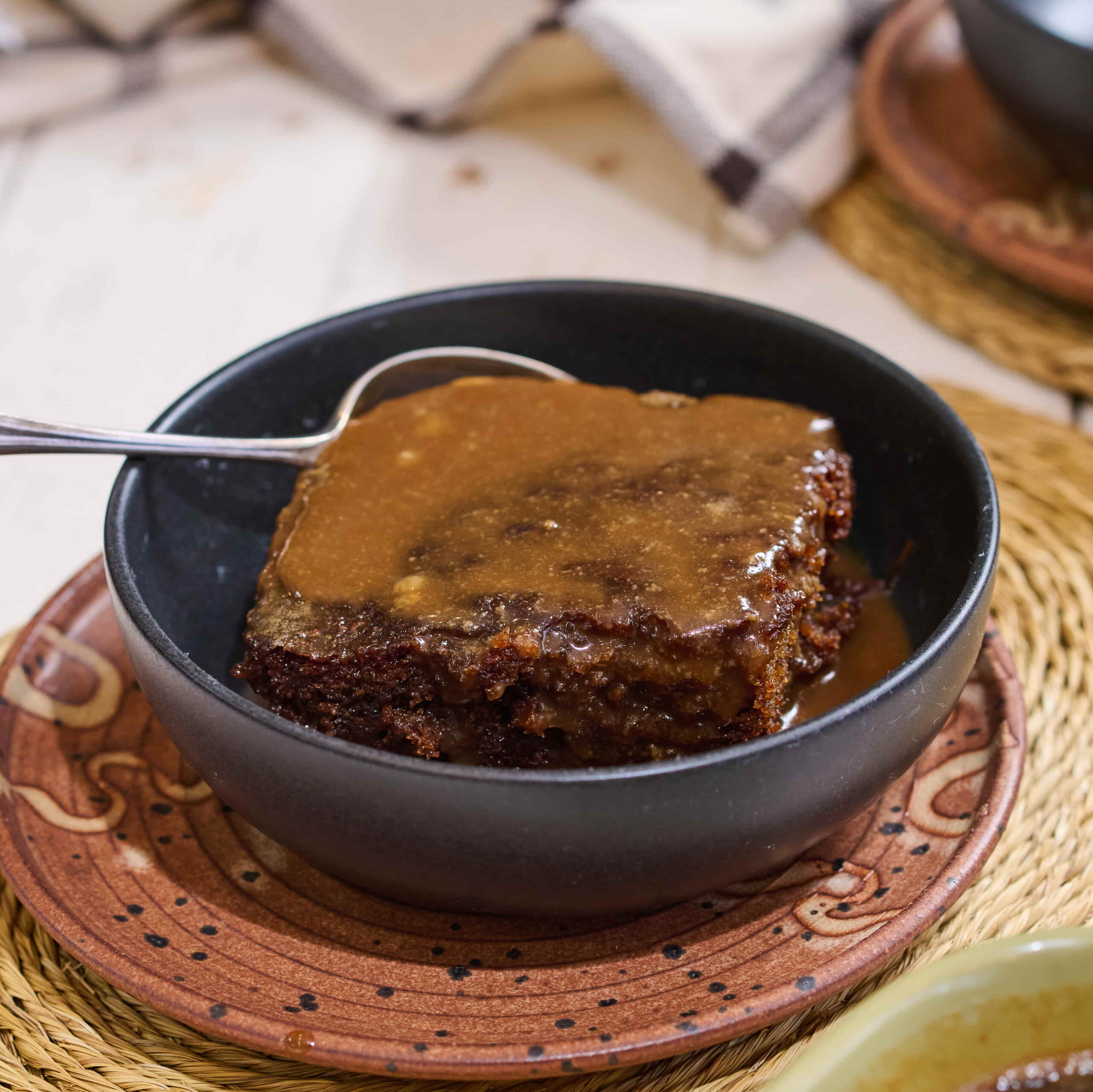 An individual portion of sticky toffee pudding covered in toffee sauce, served up in a bowl with a spoon on the side.